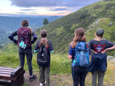 4 girls wearing backpacks facing a green hillside with clouds in the distance