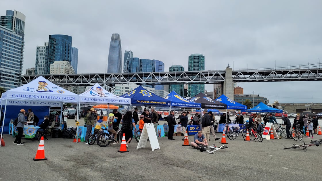 Seven popup tents with logos from san francisco area first responders. Several scouts on bikes and adults standing near the tents. Bay bridge behind.
